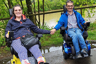 Two people in electric wheelchairs holding hands on an outdoor path, surrounded by green nature and a fence in the background.