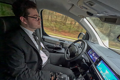 A young man in a suit independently drives a modified vehicle with joystick control on an open road.