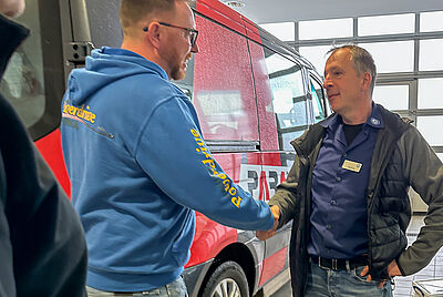 Two men greet each other with a handshake in front of a vehicle inside a workshop.