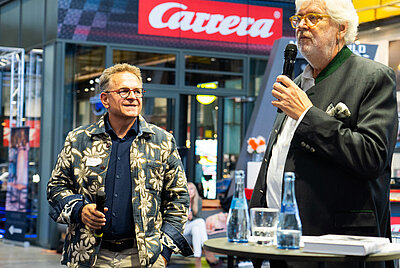 Two speakers on stage; the one on the left holding a microphone, the man on the right speaking, with “Carrera” logo behind them and audience in the background.