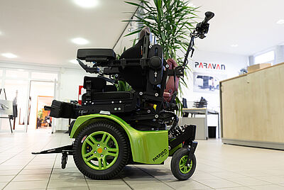 Electric wheelchair with green rims and an adjustable seating system on display in a modern showroom with the Paravan logo in the background.