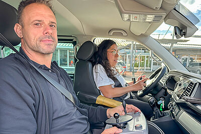 Two people inside a vehicle; the front passenger is holding an external joystick control unit while the driver operates the steering wheel.