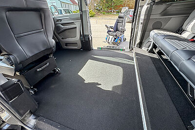 The rear interior of a VW New Caravelle, where you can look in through one side door and see the extended cassette lift with a wheelchair in front of it through the other open door. You can also see inside the car that there is no front passenger seat, and that the floor has been lowered compared to the rear bench.