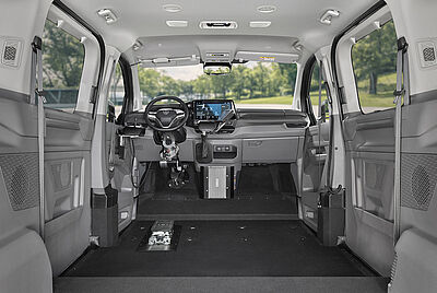 The rear interior of a VW New Caravelle, where you can look in through one side door and see the extended cassette lift with a wheelchair in front of it through the other open door. You can also see inside the car that there is no front passenger seat, and that the floor has been lowered compared to the rear bench.