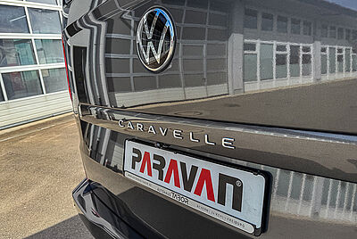 A close-up of the rear of a VW New Caravelle, clearly showing the “Caravelle” lettering and a Paravan logo in the license plate holder. The sun is shining, causing a building to be reflected in the car.