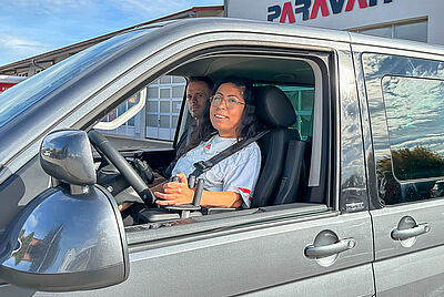 Person sitzt auf dem Fahrersitz eines umgebauten Fahrzeugs vor einem Gebäude mit dem Schriftzug „PARAVAN“; das Fahrzeug ist für barrierefreies Fahren angepasst.