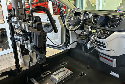 View of the vehicle interior with wheelchair restraint system in the driver's seat, joysticks for acceleration, braking, and steering, as well as head and back support