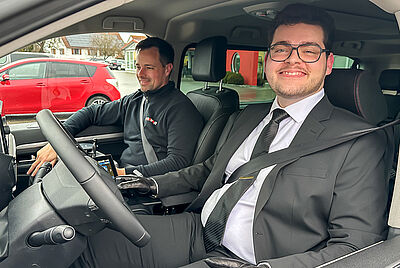 A young man in a suit sits in the passenger seat of a modified vehicle, smiling, while the PARAVAN employee looks ahead with focus.