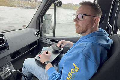 Man seated in the passenger’s seat of a van controlling the vehicle using an electronic joystick driving system.