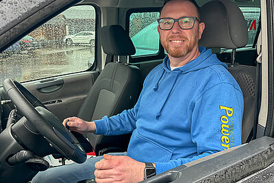 Man wearing a blue hoodie sits in the driver’s seat of a van and smiles at the camera.