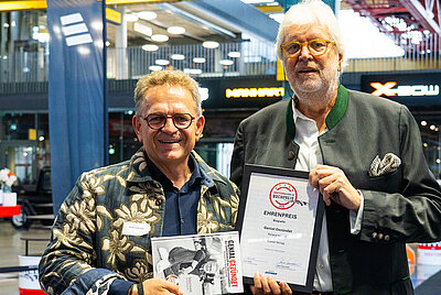 Two men in medium close-up; the man on the right presenting a certificate, the man on the left smiling and holding a book.