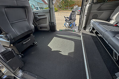 The rear interior of a VW New Caravelle, where you can look in through one side door and see the extended cassette lift with a wheelchair in front of it through the other open door. You can also see inside the car that there is no front passenger seat, and that the floor has been lowered compared to the rear bench.