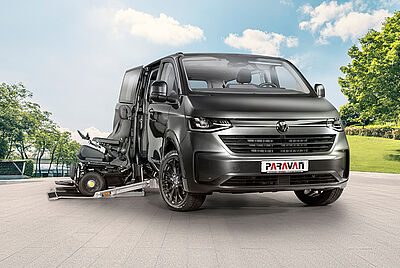 A dark gray VW New Caravelle, with its side door open, a cassette lift extended, and a PARAVAN wheelchair with a dark blue chassis parked in front of it, is parked in the courtyard of PARAVAN GmbH.