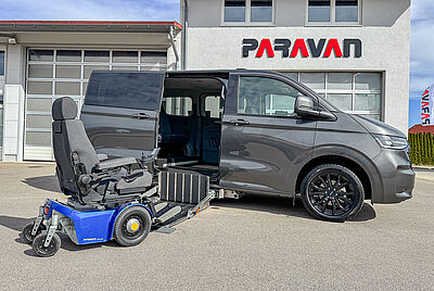A dark gray VW New Caravelle, with its side door open, a cassette lift extended, and a PARAVAN wheelchair with a dark blue chassis parked in front of it, is parked in the courtyard of PARAVAN GmbH.