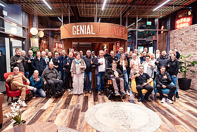 Group photo of employees and guests of Paravan GmbH in an event hall with the slogan ‘GENIAL GEZÜNDET’ displayed.
