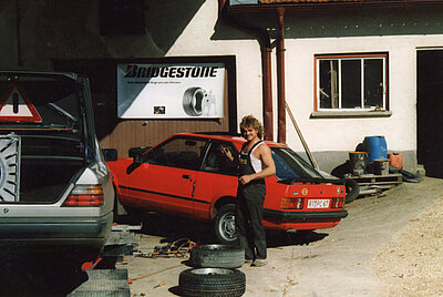 Roland Arnold working in his car workshop, standing next to a red car with an open trunk. A historic photo from PARAVAN’s early days.