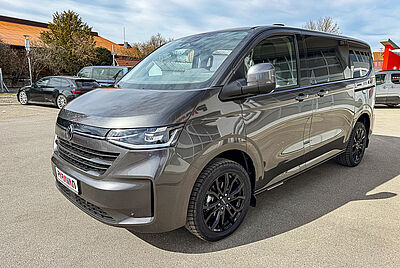 The VW New Caravelle is parked in the courtyard of the PARAVAN Mobility Park, photographed from the front right in sunny weather. The car is dark gray and has a PARAVAN logo on the front license plate holder.