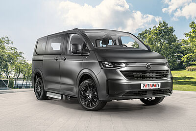 The VW New Caravelle is parked in the courtyard of the PARAVAN Mobility Park, photographed from the front right in sunny weather. The car is dark gray and has a PARAVAN logo on the front license plate holder.