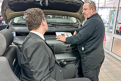 Two men discussing in front of a car with the hood open; one in a suit seated in a wheelchair, the other wearing work overalls.
