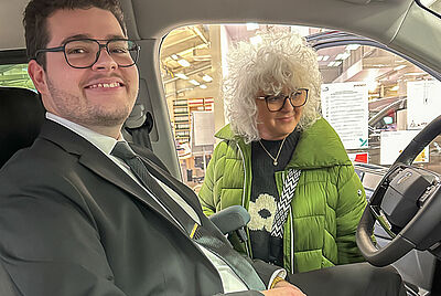 A young man in a suit smiles while seated in a modified vehicle, speaking with a curly-haired woman in a green jacket standing outside the open door.