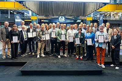 Large group of about 25 people on stage, most holding certificates; group photo at an award ceremony in a modern event venue.