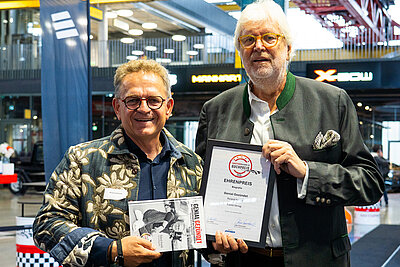 Two men standing side by side at an event hall; the man on the right holds a certificate (“Honorary Prize Biography”) and the man on the left holds a book titled “Genial Gezündet”.