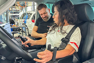 Sabina seated in the driver’s seat of a modified vehicle, operating an adapted steering system with additional grips and controls.