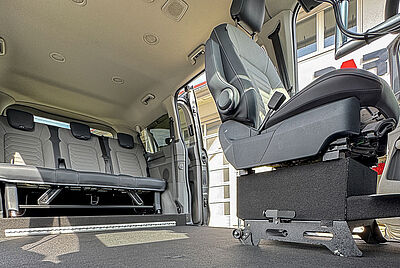 The interior of a vehicle as seen from the floor on the passenger side, where the passenger seat is no longer visible. The driver's seat, on the right in the picture, is not a standard seat but one that can be removed and moved. On the left, you can see into the back of the car, where you can see the rear bench, how much space there is inside the car, and the lowered floor.