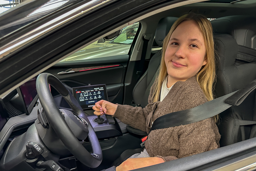 A smiling young woman with blonde hair is sitting in the driver's seat of a car, looking out the open window. She is holding a joystick in her right hand. 