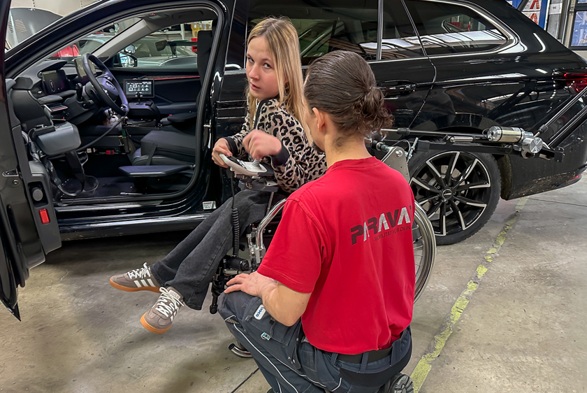 A young blonde woman is sitting in her wheelchair, parallel to a black car with the driver's door open. A robotic arm extends from the trunk toward the wheelchair on the right side of the picture. A man wearing a red Paravan T-shirt is kneeling in front of her. 