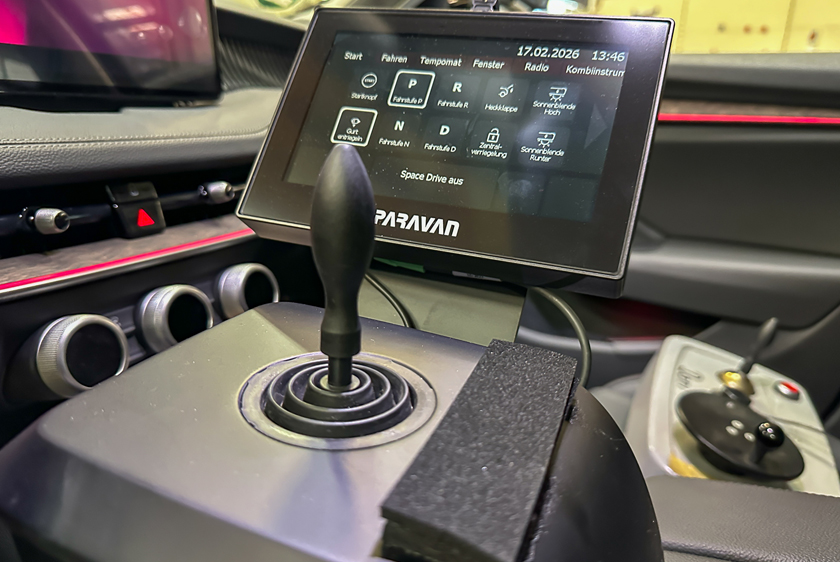 In the foreground, you can see a joystick installed in a car. Behind the joystick is a Paravan touchscreen display showing various driving functions. 