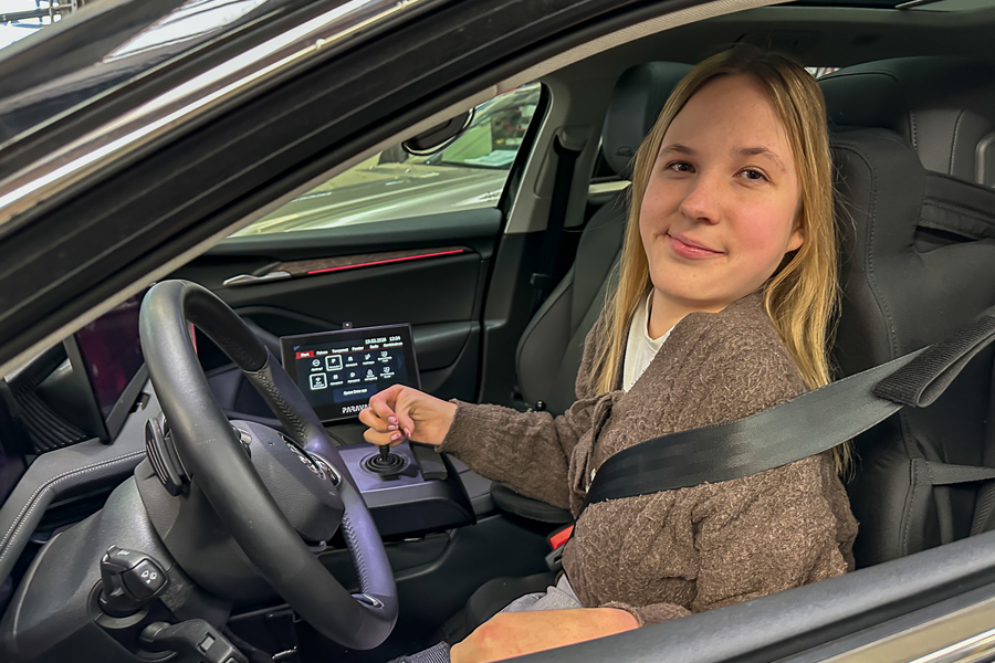 A smiling young woman with blonde hair is sitting in the driver's seat of a car, looking out the open window. She is holding a joystick in her right hand. 