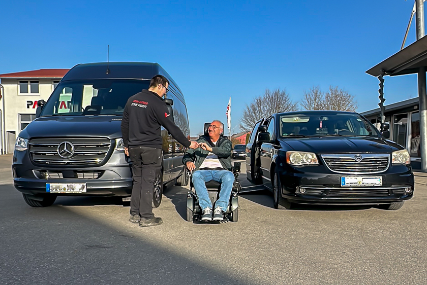 Under a blue sky, two vehicles are parked in the Paravan courtyard: a new Sprinter on the left and a Chrysler Voyager on the right. Between the two vehicles, a man is sitting in a wheelchair, smiling, as a Paravan employee shakes his hand.