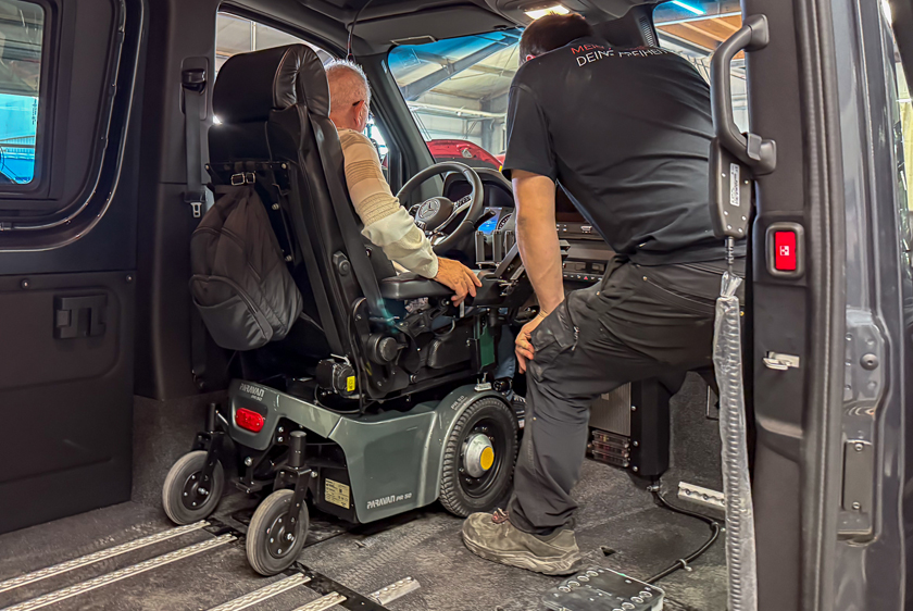 A man in a Paravan PR50 electric wheelchair is standing in front of the steering wheel inside a Sprinter.  In place of the passenger seat, there is a Paravan employee looking on intently.
