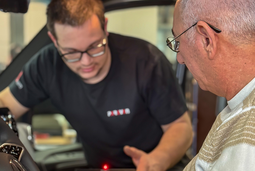 In the foreground, on the left, is a car steering wheel, and on the right is a man with short white hair, glasses, and a brown striped sweater. In the background, out of focus, a Paravan employee is explaining the Paravan Touch Display.