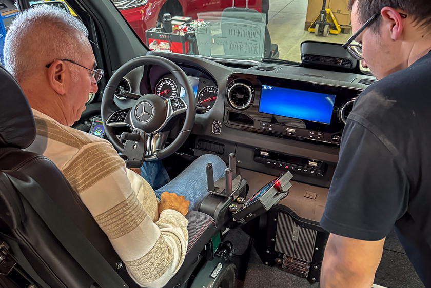 A man in a wheelchair is sitting in the driver's seat of a car that has been modified with alternative controls, such as a trident on the steering wheel. On the far right, partially in the frame, another man is standing and looking down.