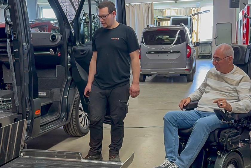 To the right of a vehicle, a man in a wheelchair is waiting. Another man is standing closer to the vehicle; both are looking at the cassette lift that has just been extended and is mounted on the car. 