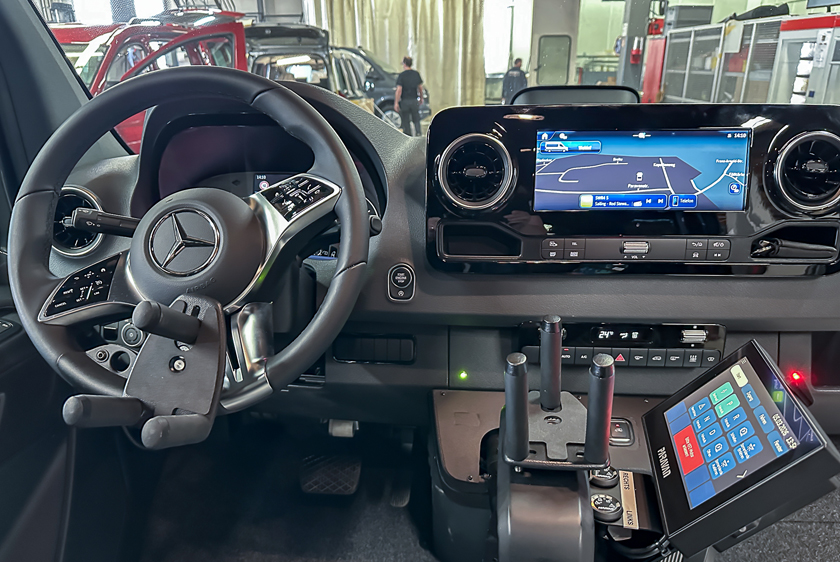 On the left, you can see a car steering wheel fitted with a trident. To the right of the steering wheel is another trident joystick, installed as an accelerator and brake control. The Paravan Touch Display was mounted on it. 