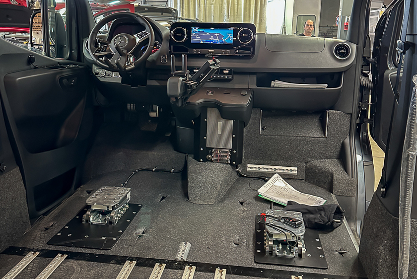 The interior of a car that has undergone extensive modifications. In addition to changes made to the steering wheel for controlling the vehicle, both the driver's seat and the passenger seat have been removed and replaced with a docking station. 