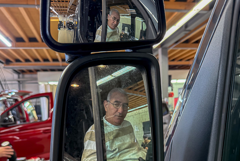 A vehicle's side mirror. The reflection of a man wearing glasses and a brown striped sweater can be seen. 