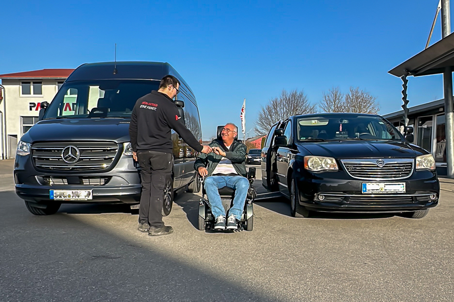 Under a blue sky, two vehicles are parked in the Paravan courtyard: a new Sprinter on the left and a Chrysler Voyager on the right. Between the two vehicles, a man is sitting in a wheelchair, smiling, as a Paravan employee shakes his hand.
