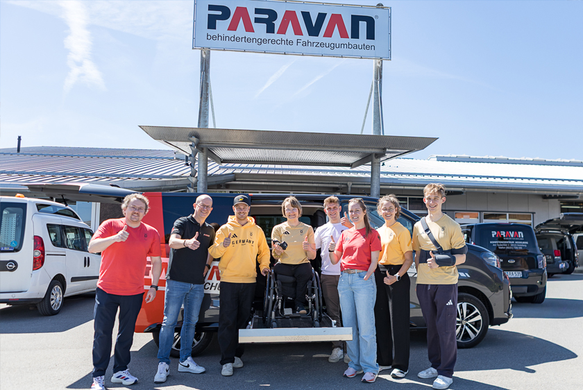 Several people are standing in front of a car in bright sunshine outside the Paravan Mobility Park. A woman in a wheelchair is standing on the extended cassette lift, high up so she’s the same height as everyone else. Except for two, everyone in the picture is wearing either red or yellow Team Germany clothing. 