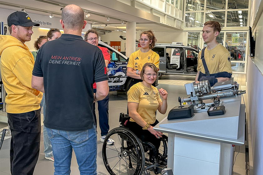 Several people are standing in a loose circle while an employee of Paravan GmbH explains something to them. To the right is a display table with joysticks...