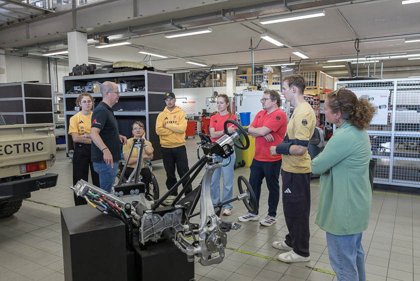 In a workshop, a group of about eight people is standing in front of a display that shows a vehicle axle connected to the steering wheel. An employee is explaining how it works.