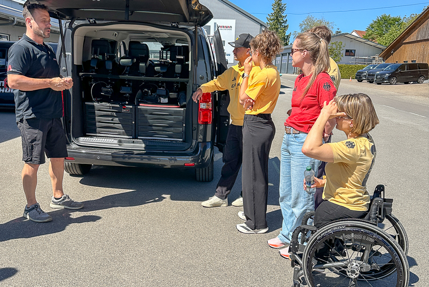 On the left, the driving instructor from Paravan Driving School stands in front of a vehicle with its trunk open, inside which are several joysticks. To the right of the trunk, six people are standing in Team Germany uniforms. All of this is taking place under bright sunshine. 