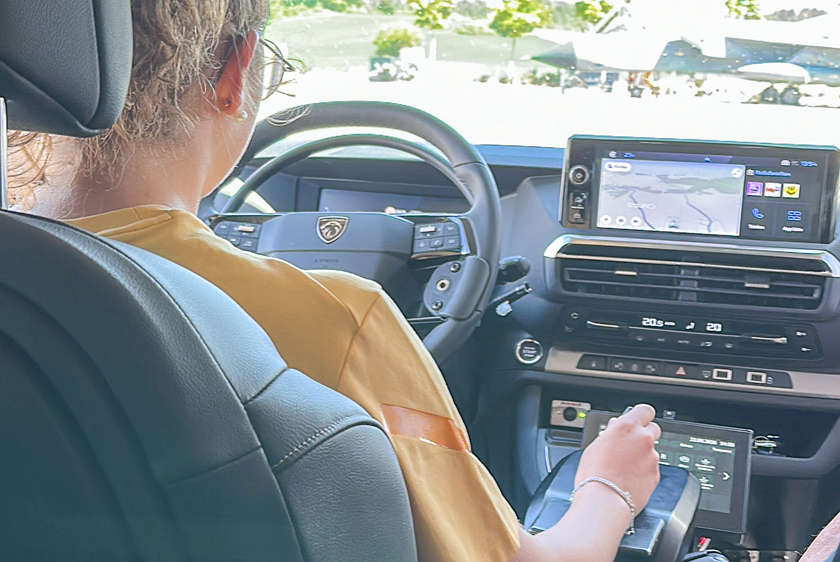 A woman, seen from over her right shoulder, is driving a car using joysticks.