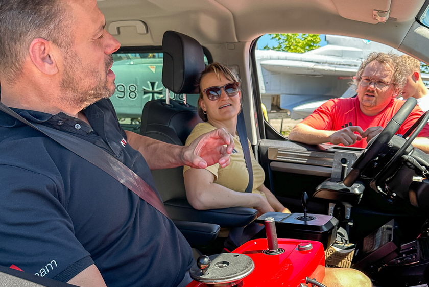 Through the open passenger-side window, you can see two people sitting in the vehicle, as well as another person looking in through the open driver-side window. The Paravan Driving School instructor is sitting in the passenger seat, and a woman wearing sunglasses is in the driver's seat. All three people are talking to each other.