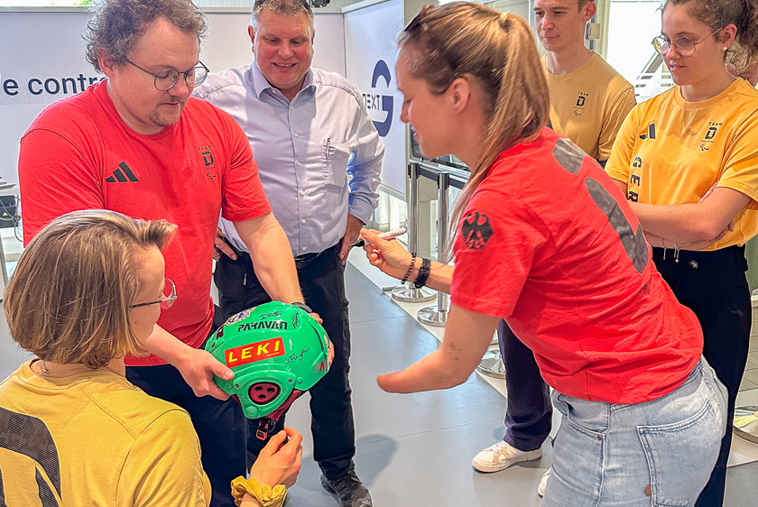 Six people are standing in a circle. Everyone is looking at a green LEKI helmet that the people in the circle are signing. 