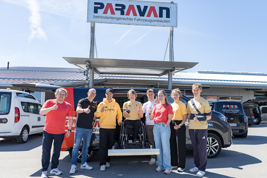 Several people are standing in front of a car in bright sunshine outside the Paravan Mobility Park. A woman in a wheelchair is standing on the extended cassette lift, high up so she’s the same height as everyone else. Except for two, everyone in the picture is wearing either red or yellow Team Germany clothing. 