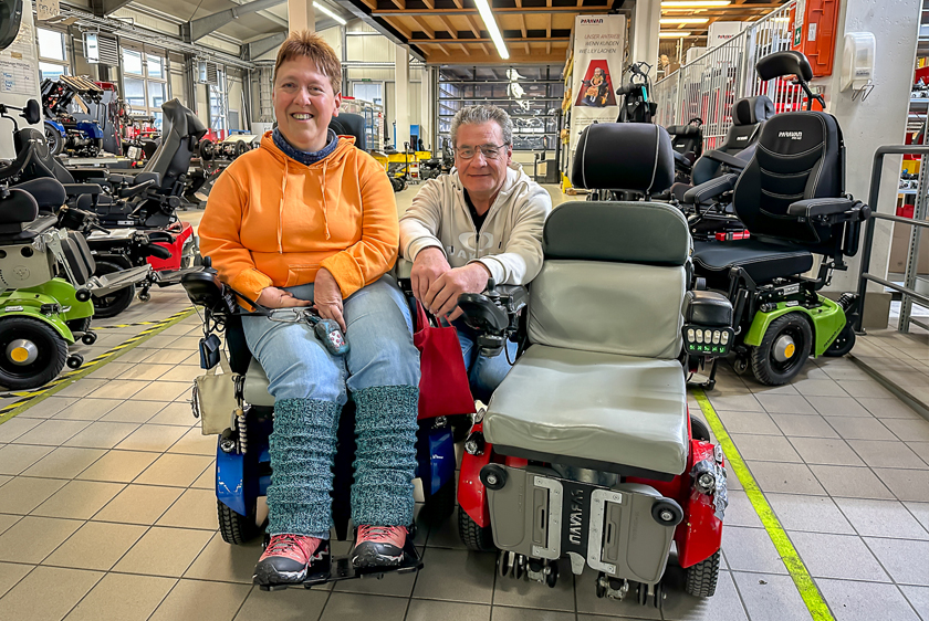 A woman wearing an orange sweater and sitting in a new blue wheelchair, with an older red wheelchair to her right, is standing in the wheelchair workshop at Paravan GmbH. The head of the wheelchair department is kneeling in the middle, behind the two wheelchairs. 